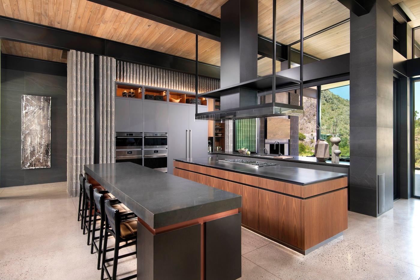 Spacious modern kitchen with sleek black countertops, wooden cabinetry, and large windows. Bright natural light reveals mountain views, creating an open, airy feel.