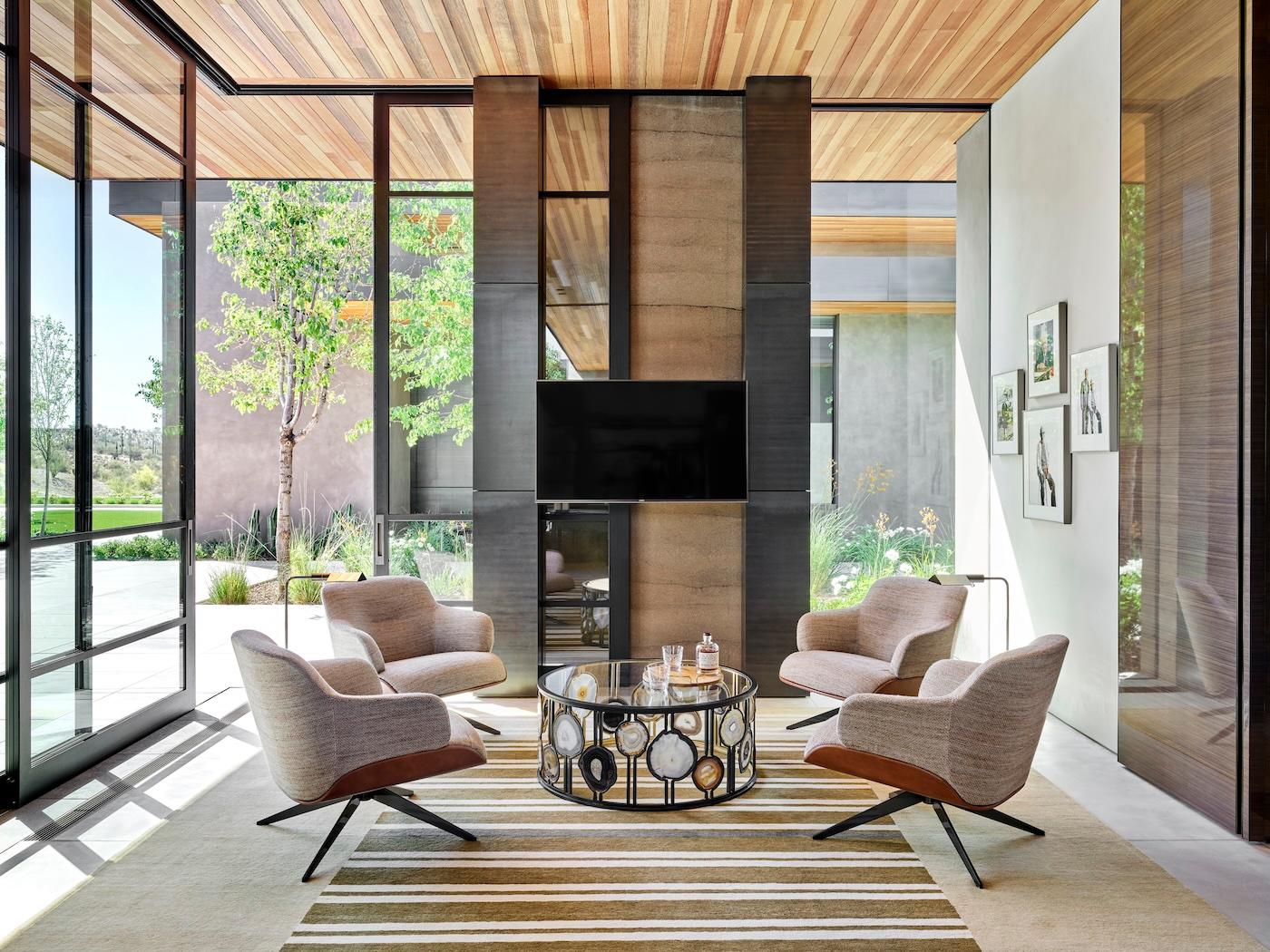 Modern living room with large windows and natural light. Four beige swivel chairs surround a glass coffee table on a striped rug; serene and inviting ambiance.