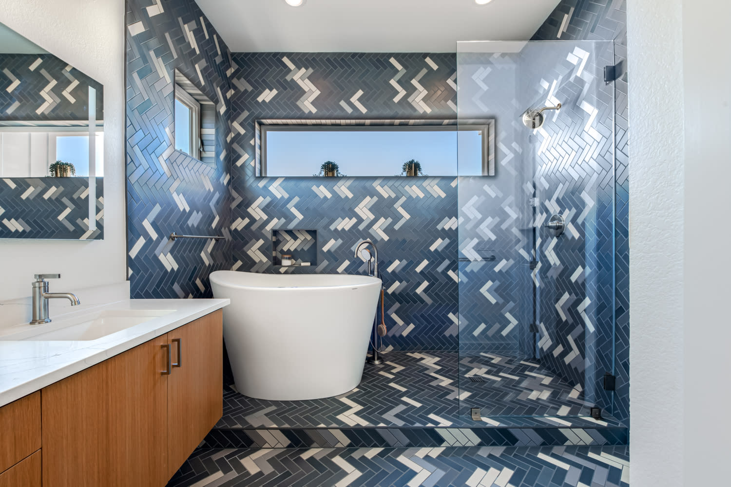 This bold bathroom showcases striking blue herringbone tiles accented with white, creating a dynamic backdrop for the freestanding tub and glass-enclosed shower. Warm wood cabinetry and modern fixtures complete the contemporary, spa-like retreat.