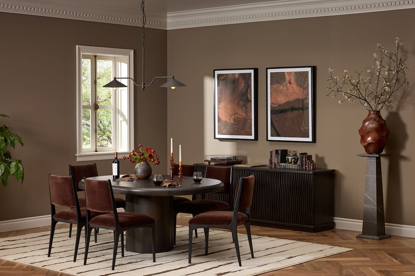 Sophisticated dining room with a dark round table, four brown chairs, and a modern chandelier. Earth-toned art, sideboard, and a vase add elegance.