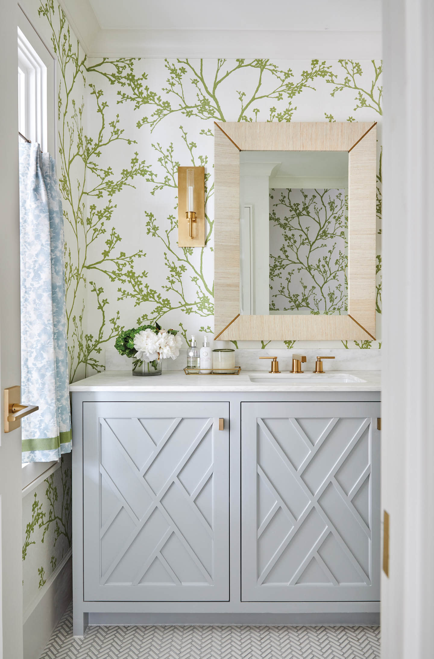 Airy bathroom with pale botanical wallpaper, blue vanity, faceted mirror, and sheer floral curtain.