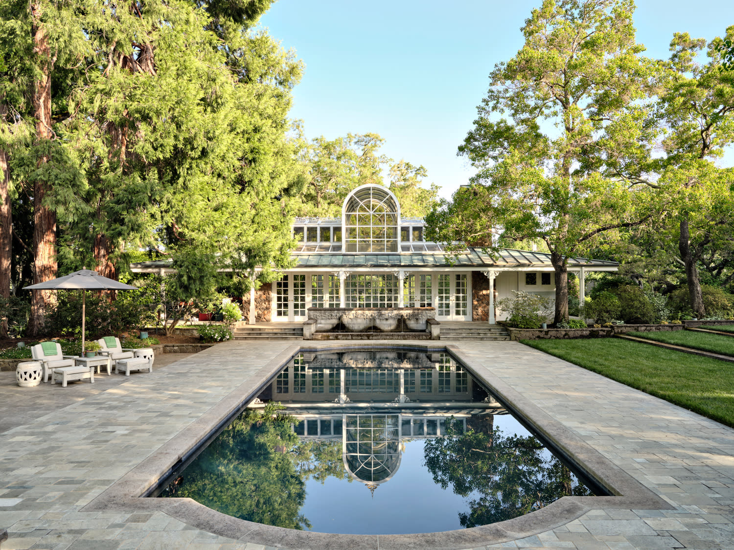Elegant pool house with arched windows, surrounded by lush trees.
