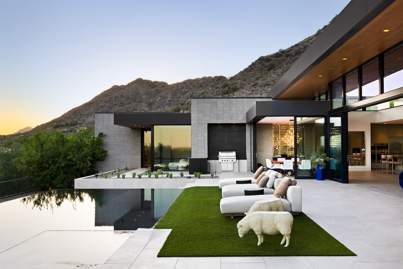 Modern luxury home with sleek architecture and large windows, features an outdoor patio with white lounge chairs, green turf, and a pool. Mountain backdrop at sunset.
