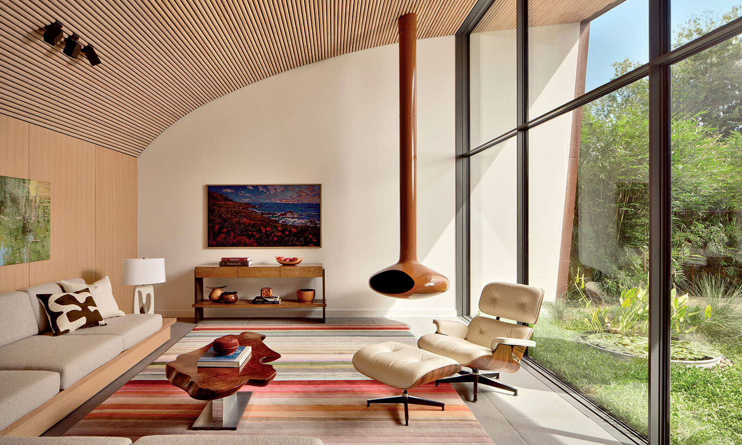 Modern living room with high wood-paneled ceiling, large windows, natural light, beige sofa, Eames lounge chair, hanging fireplace, and colorful striped rug.