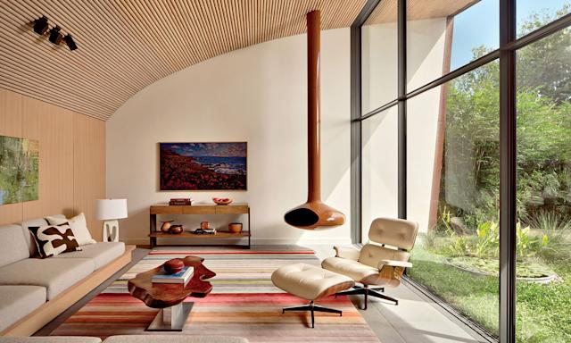 Modern living room with high wood-paneled ceiling, large windows, natural light, beige sofa, Eames lounge chair, hanging fireplace, and colorful striped rug.