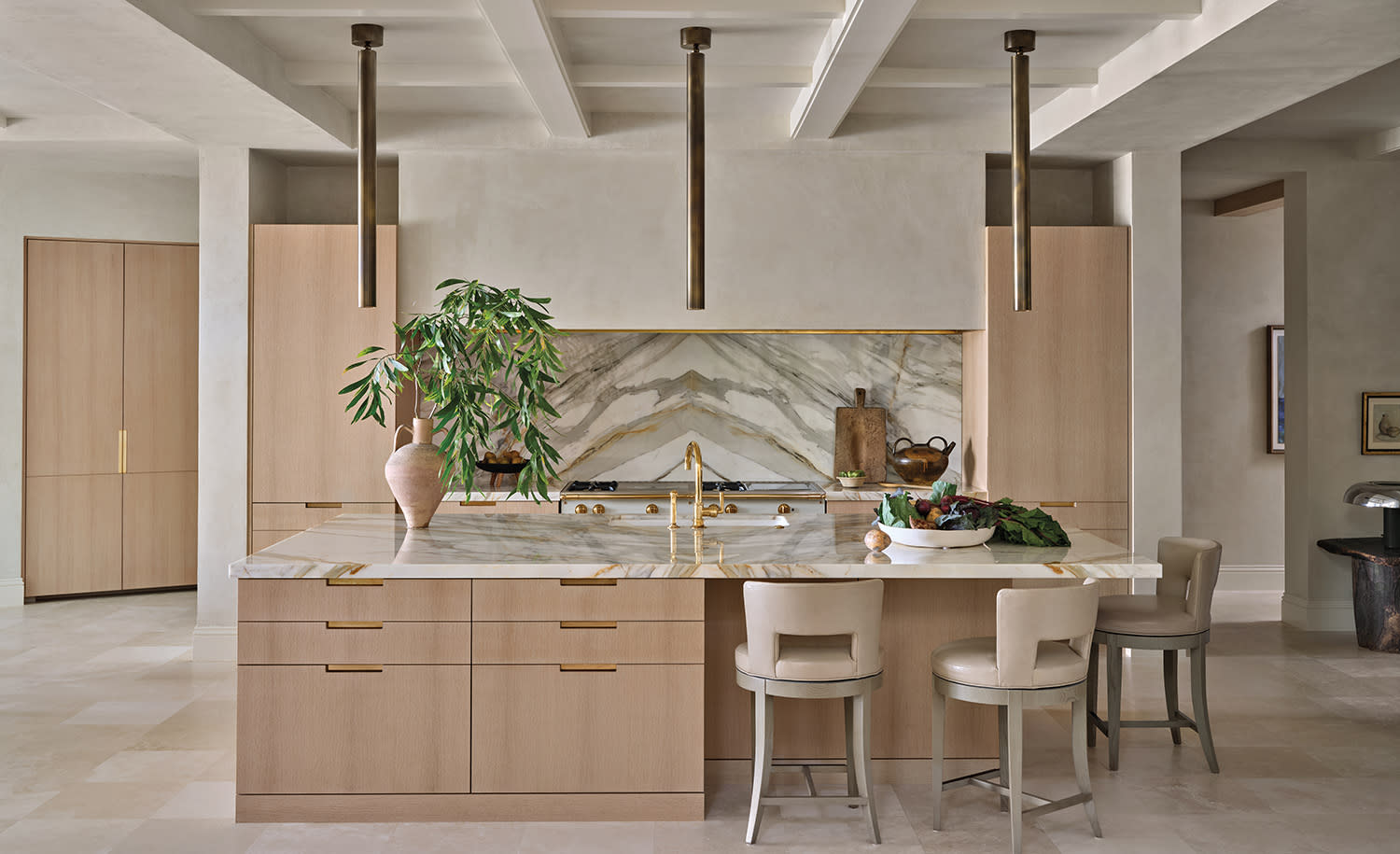 A modern kitchen with neutral tones features a large marble island, gold fixtures, and hanging lights. 