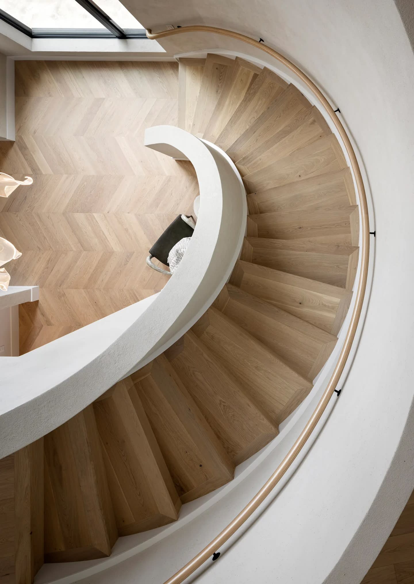 Curved wooden staircase with light wood steps viewed from above. It spirals around a central column, leading down to a chevron-patterned wood floor.