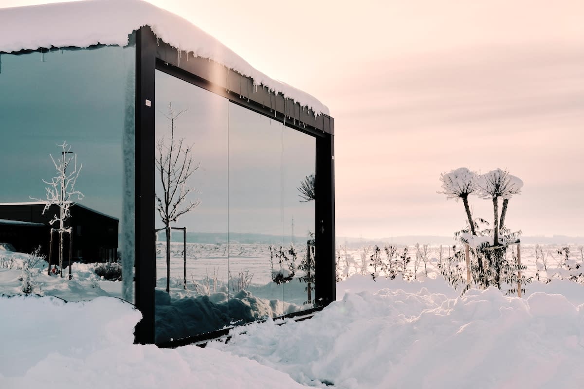 Glass-enclosed pergola structure surrounded by snow, with frosted trees and a winter landscape reflected in the transparent walls.