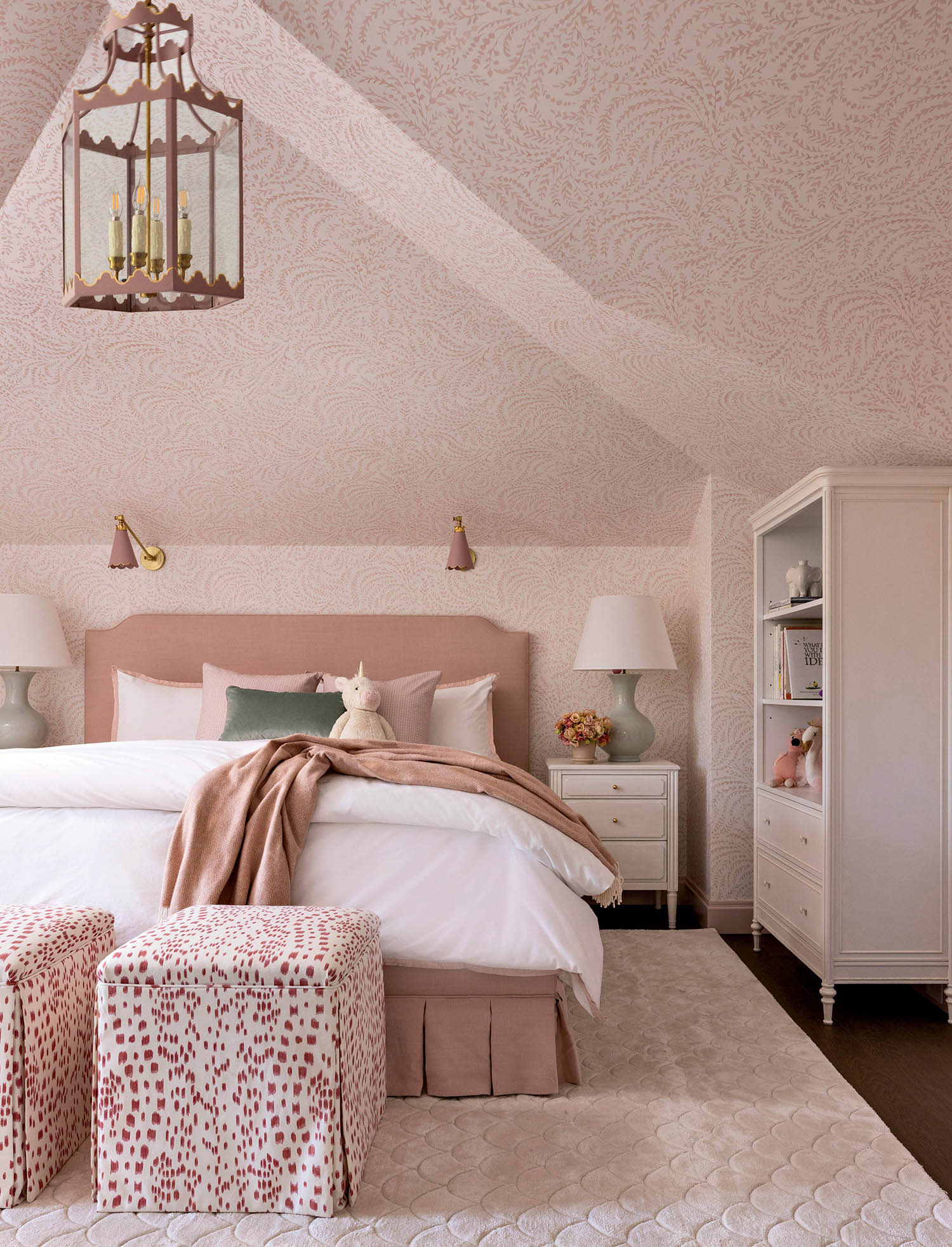 A bedroom decorated in pink wallpaper, a matching rug and patterned ottomans, along with a bookcase.