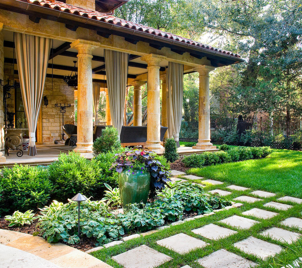 Decorative stone path on well manicured lawn lead to spacious veranda with concrete pillars and curtains for privacy.