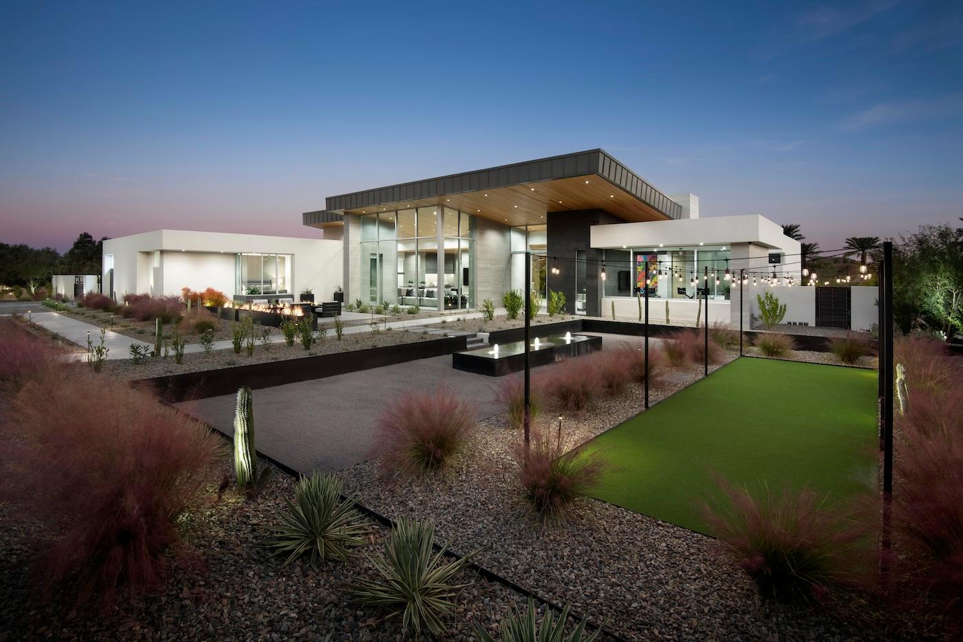 Modern house at dusk with sleek, angular design. Large glass windows, manicured lawn, desert landscaping, and string lights create an elegant atmosphere.