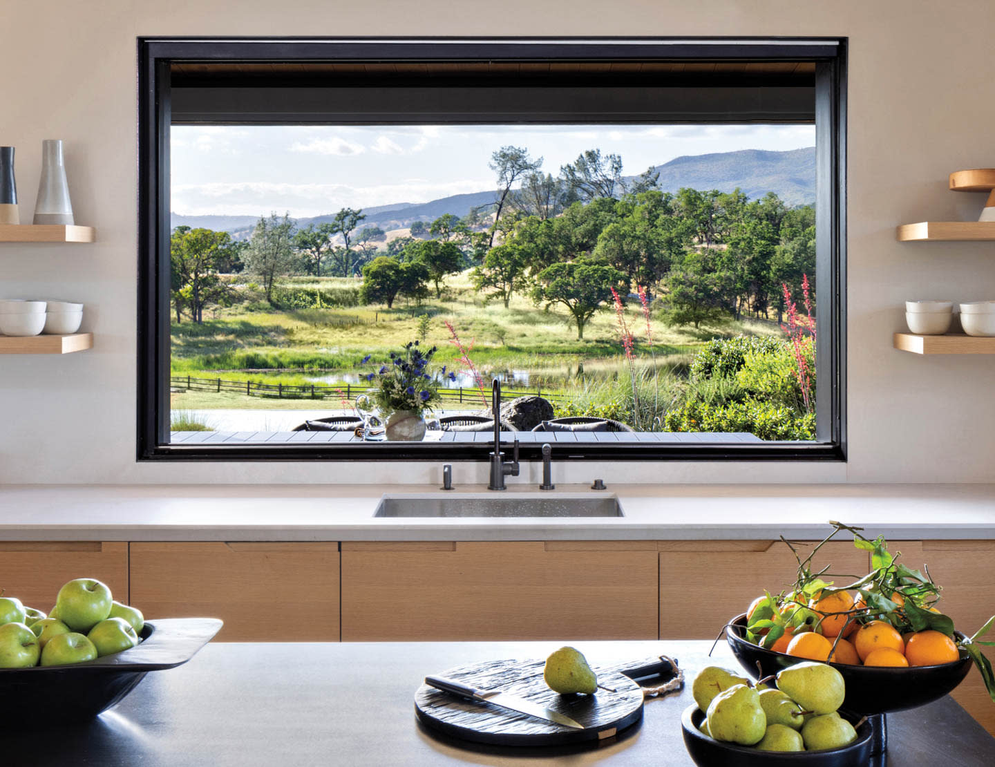 Modern kitchen with large window overlooking lush green landscape and hills. Fresh fruit bowls on countertop, creating a serene, inviting atmosphere.