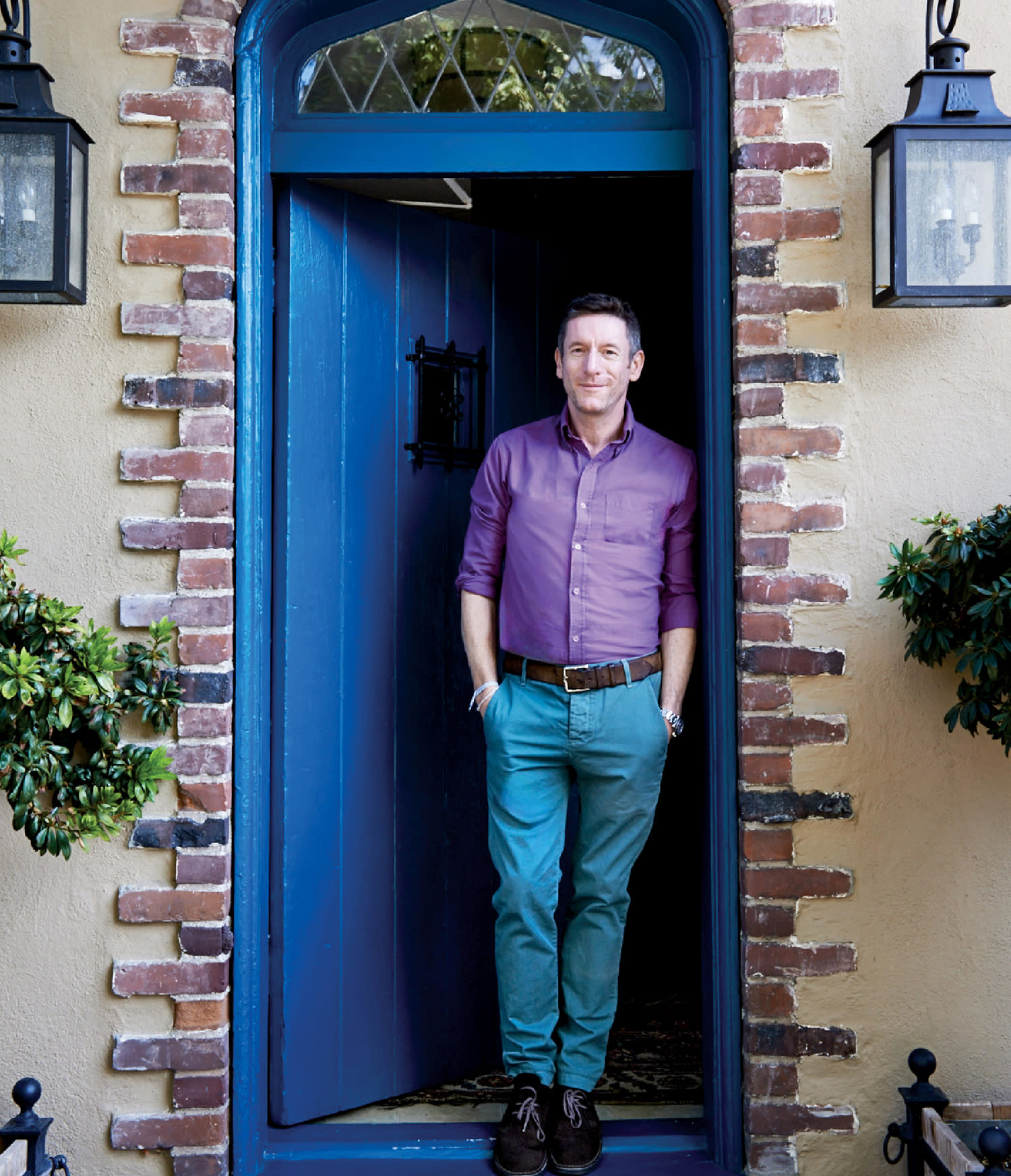 Designer Peter Dunham standing in a blue doorway framed by brick and lantern-style lights