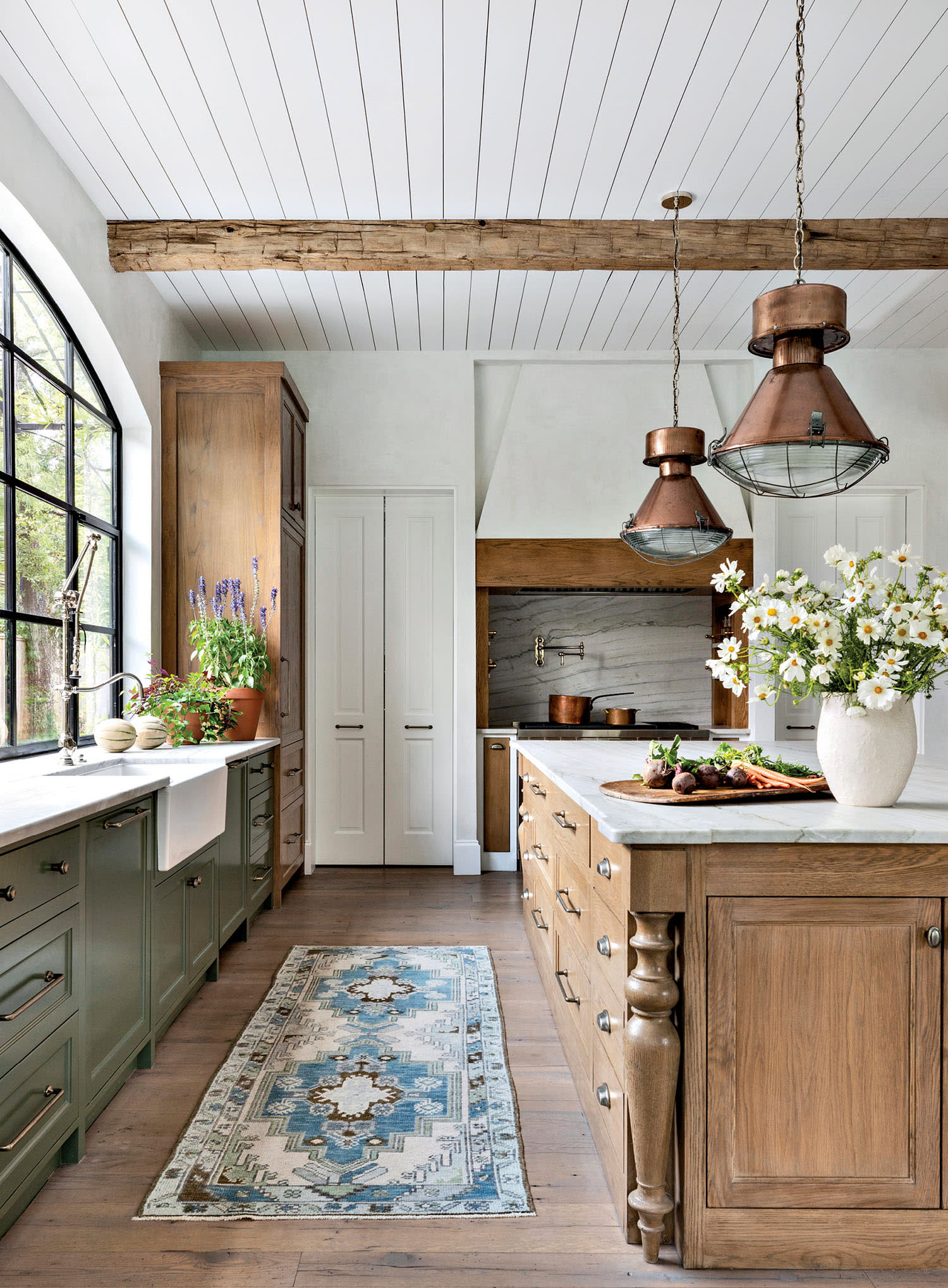 A rustic kitchen features a wooden island with a vase of daisies, copper pendant lights, and green cabinets.