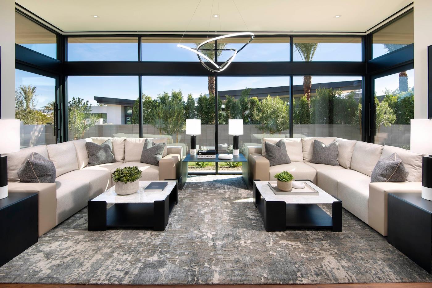 Modern living room with floor-to-ceiling windows, beige sofas, black coffee tables, and geometric chandelier. Natural light and greenery add a serene vibe.