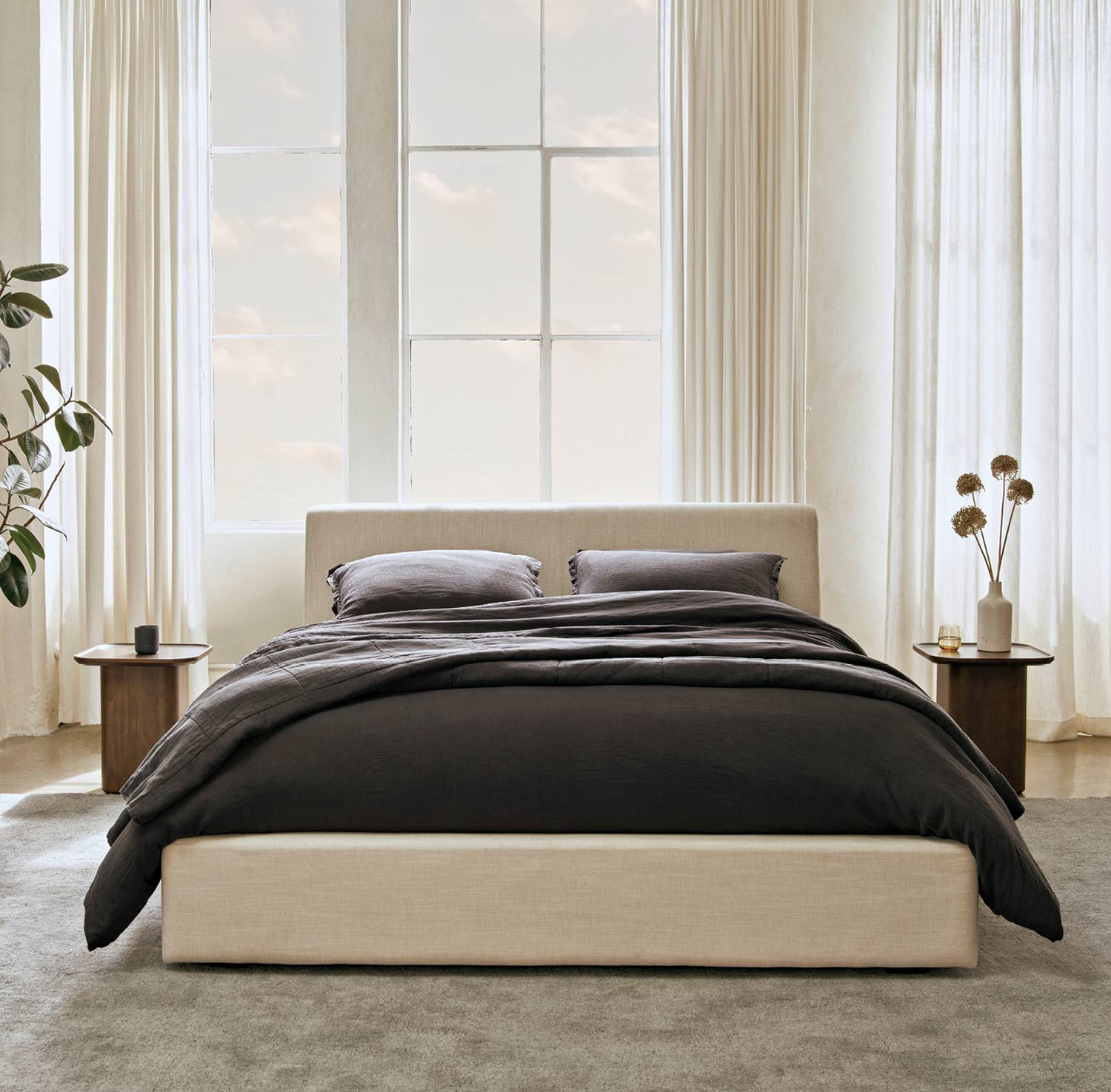 A bed made up with dark linens in a room with white curtains.