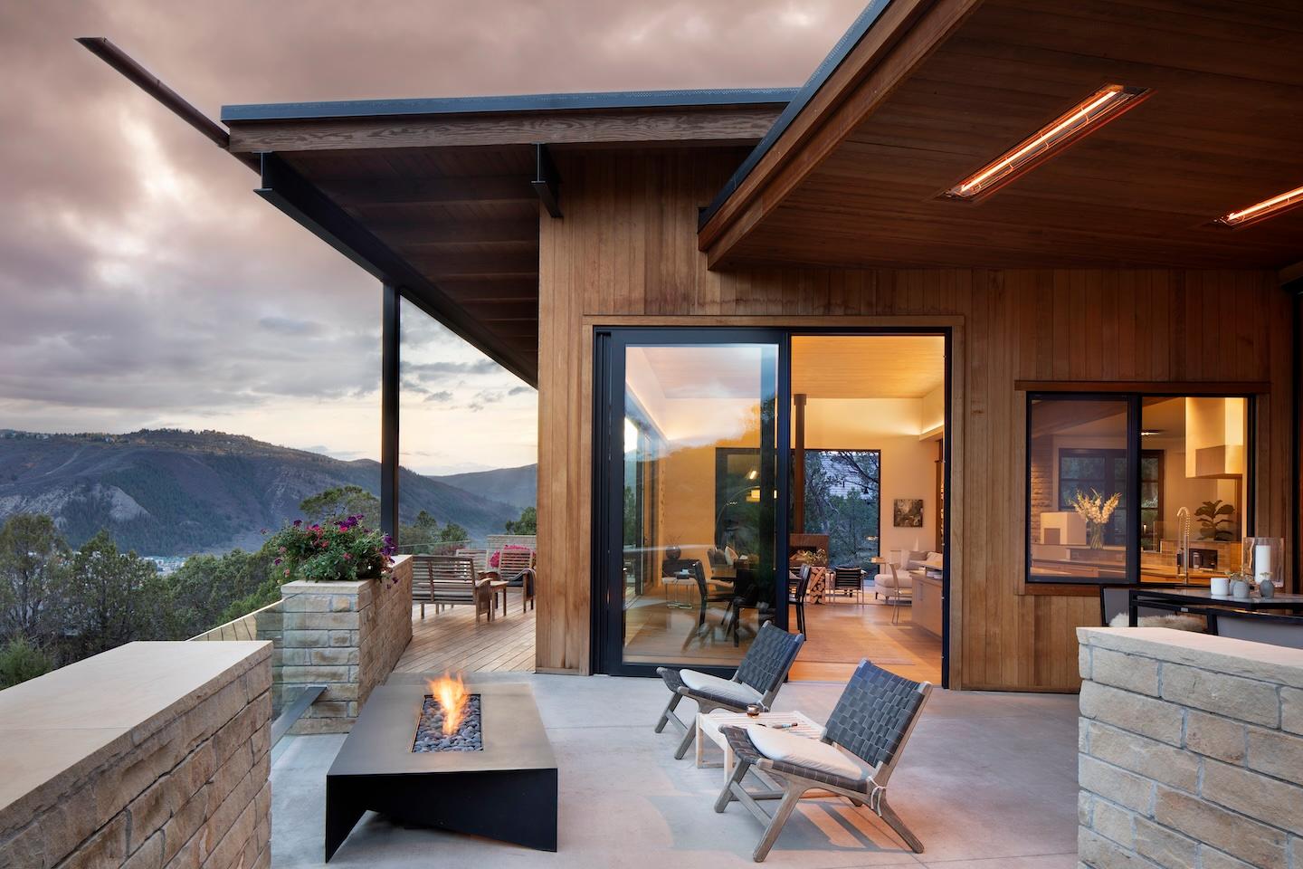 Modern wooden house with mountain views at dusk. The patio features a fire pit and seating, creating a cozy atmosphere. Indoor lights illuminate the open interior.