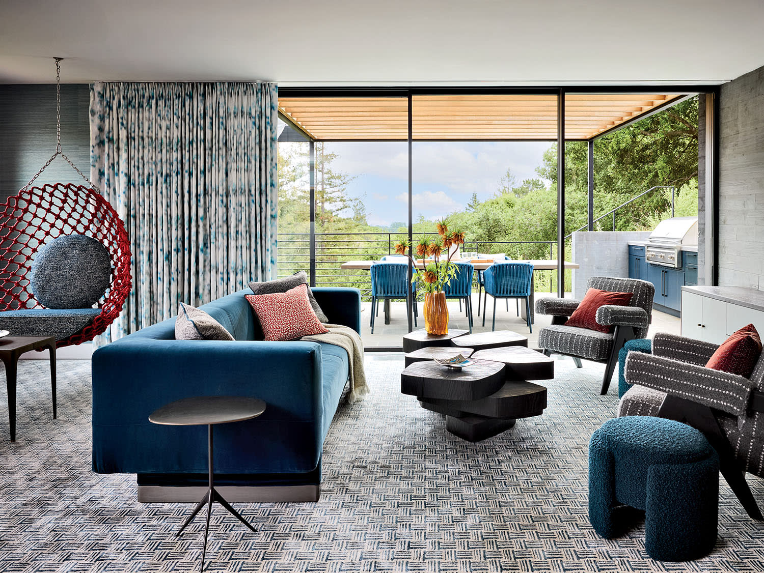 Modern living room with a blue sofa, patterned carpet, and red hanging chair. Glass doors reveal a patio with lush greenery, creating a serene atmosphere.