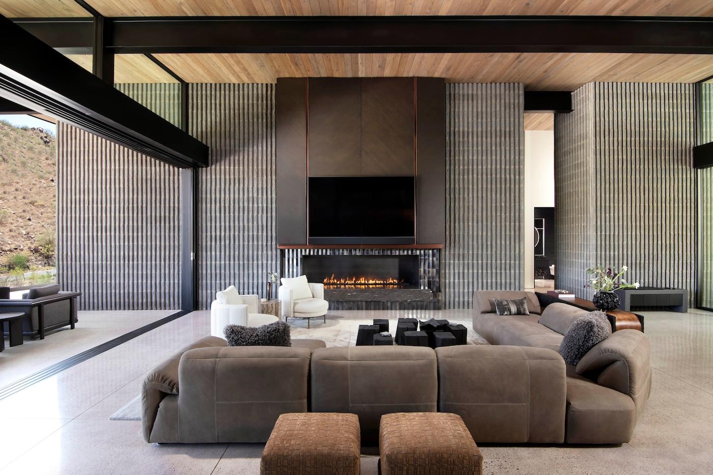 Spacious modern living room with high ceilings, large gray sectional sofa, fireplace under a wall-mounted TV, and natural light from expansive windows.