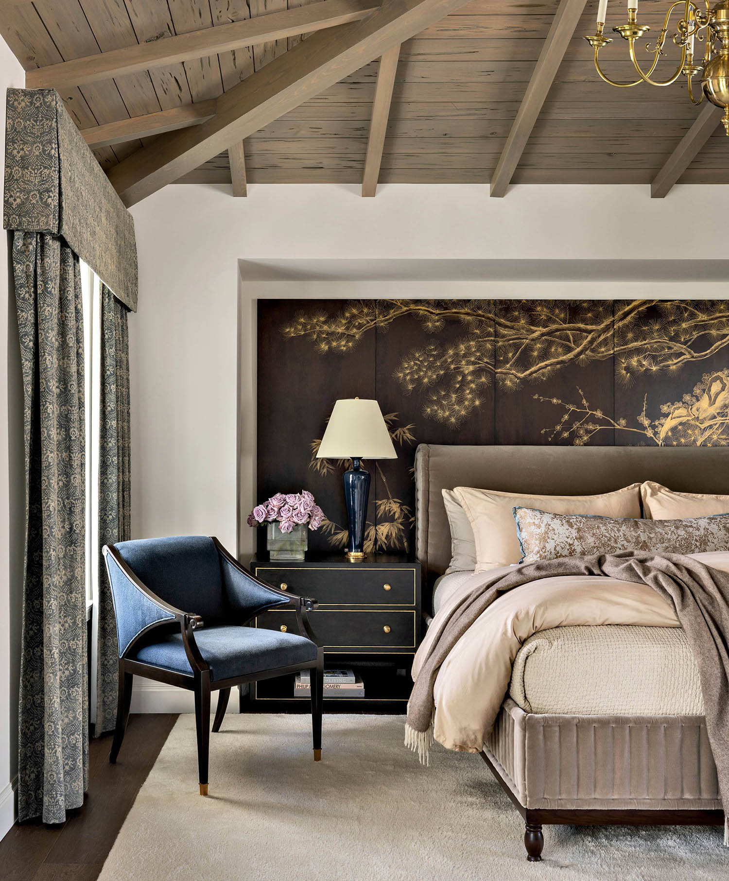 A bed and a blue chair sit in a bedroom with wood-paneled ceilings, a nightstand and curtained window.