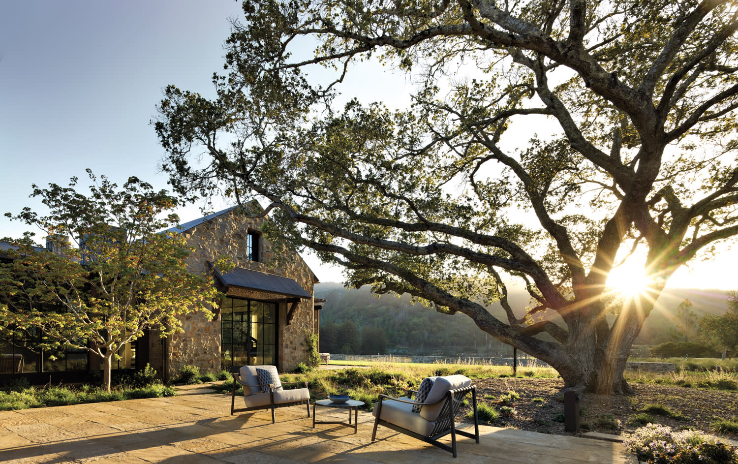 Exterior of a large home with black chairs and table, large oak tree and view of the vineyard and mountains