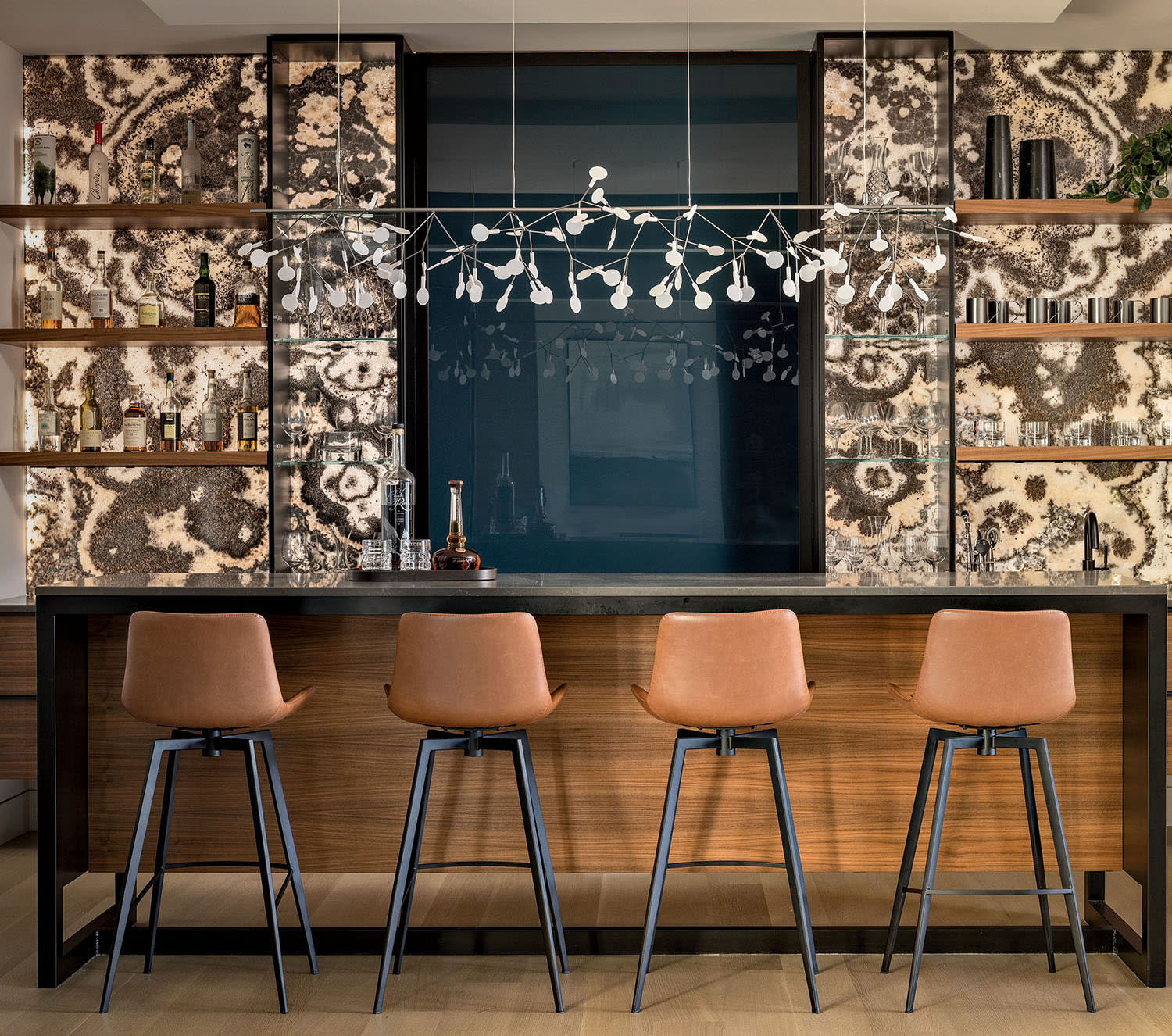 A wood bar with shelves sits near a patterned wall, with leather stools lined up at the edge.