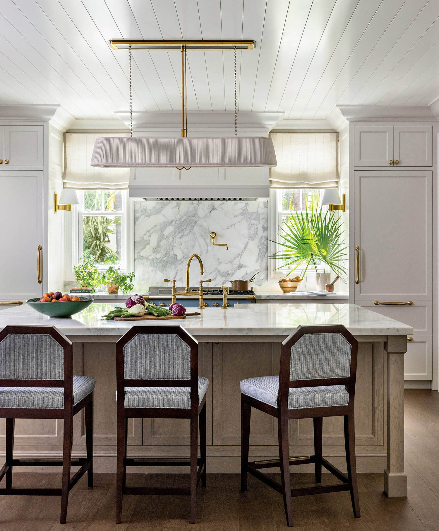 A white kitchen with a center island lined with stools, bright windows and an overhead chandelier.