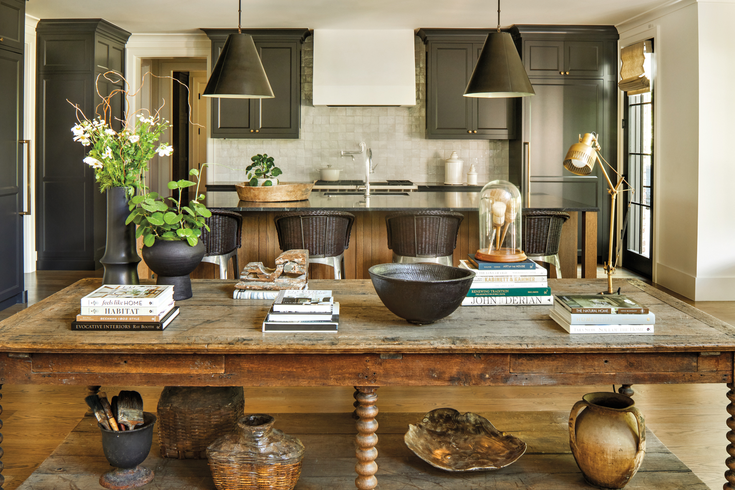 antique wooden table backed by a kitchen with black cabinetry and pendant lights over an island