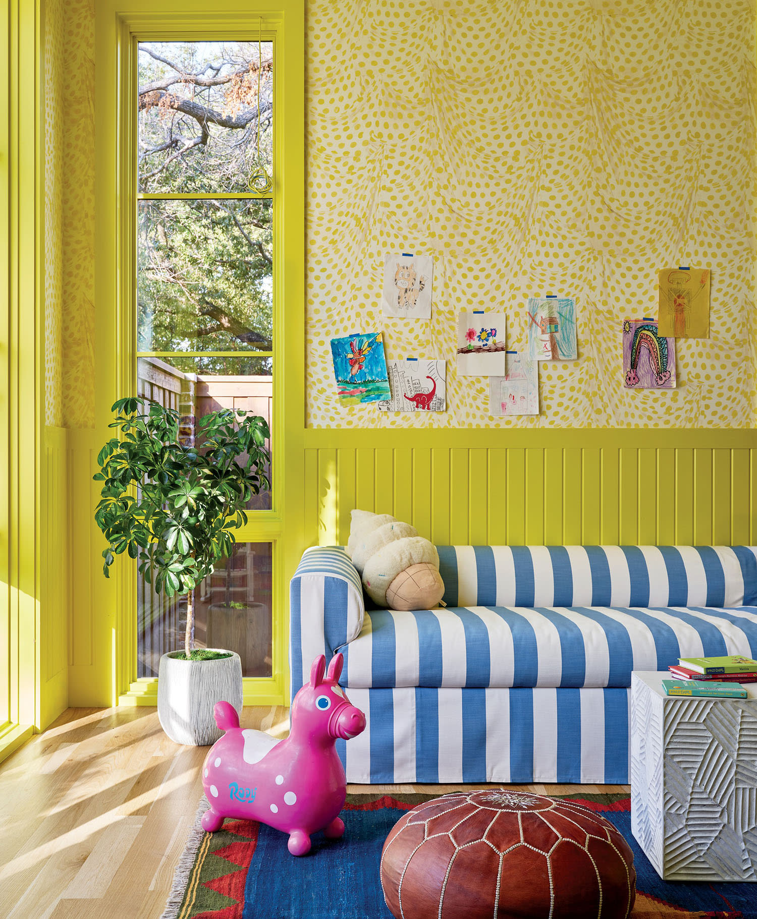 Bright, playful room with yellow walls and large window. Blue-striped sofa, kids' drawings on wall, pink inflatable toy, plant, and brown pouf create a cheerful vibe.