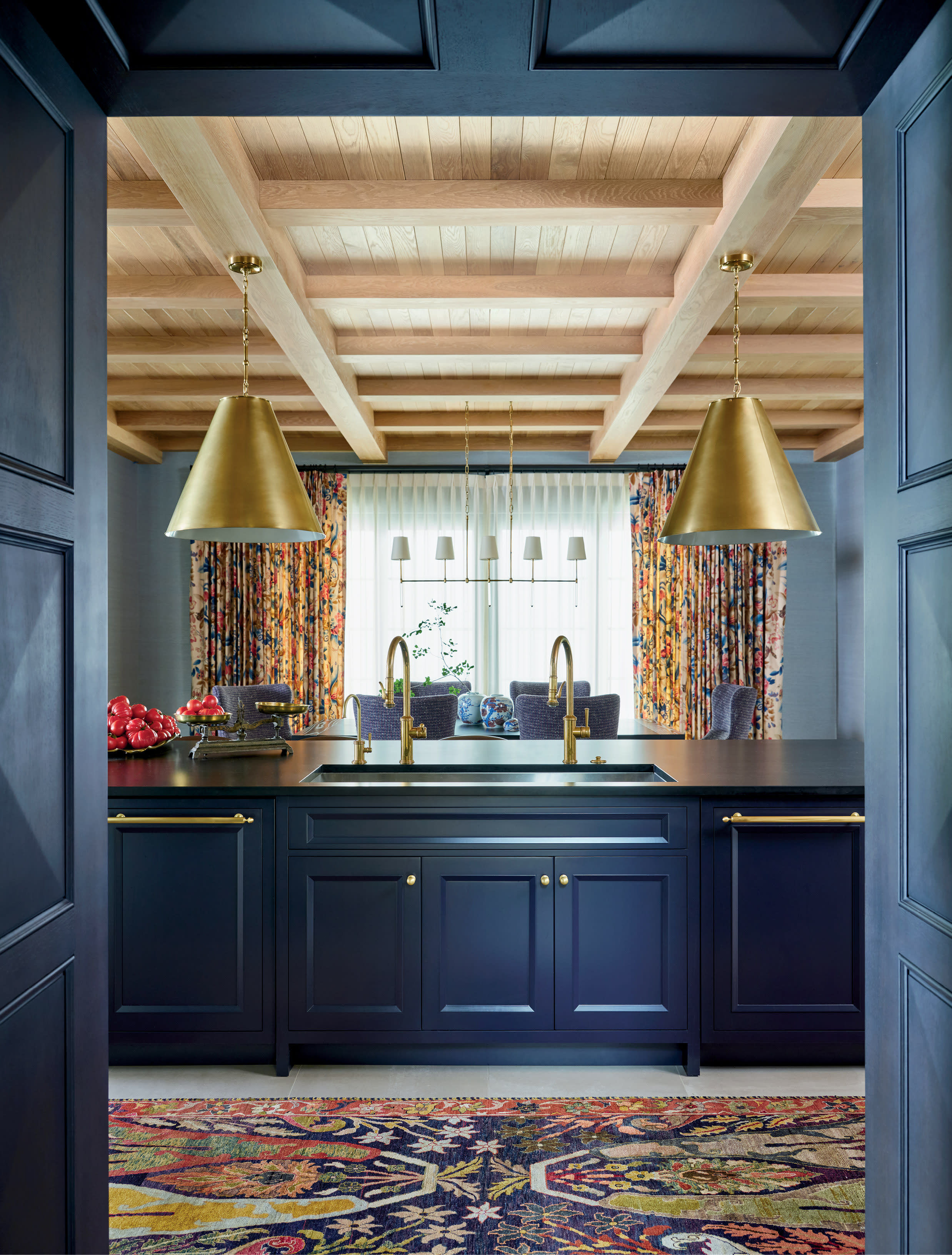 Elegant kitchen with navy cabinets, gold fixtures, and two large brass pendant lights. A colorful rug and vibrant curtains add warmth against a wooden ceiling.