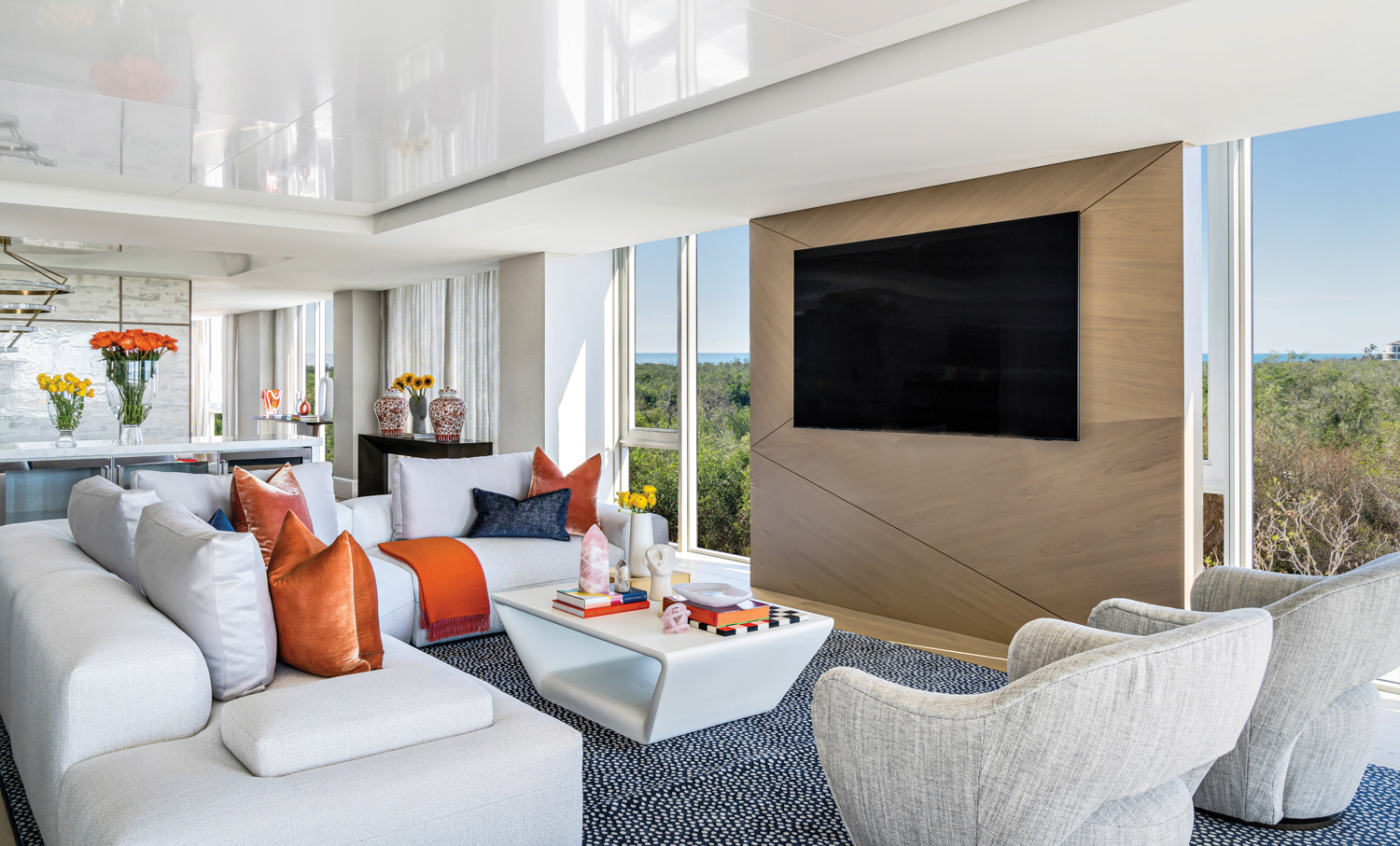 living area with blue spotted rug, white sectional and coffee table, gray chairs, wood television wall and orange pillows