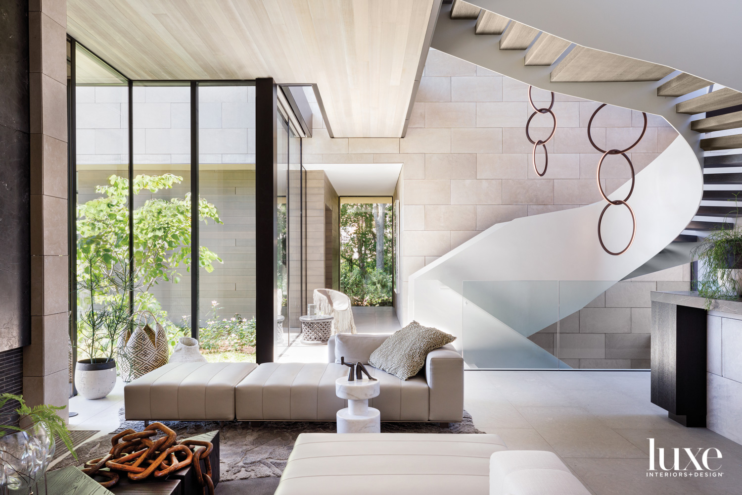 A living room with floor-to-ceiling windows and a modern curving white staircase. Circular pendant lights hang from above.