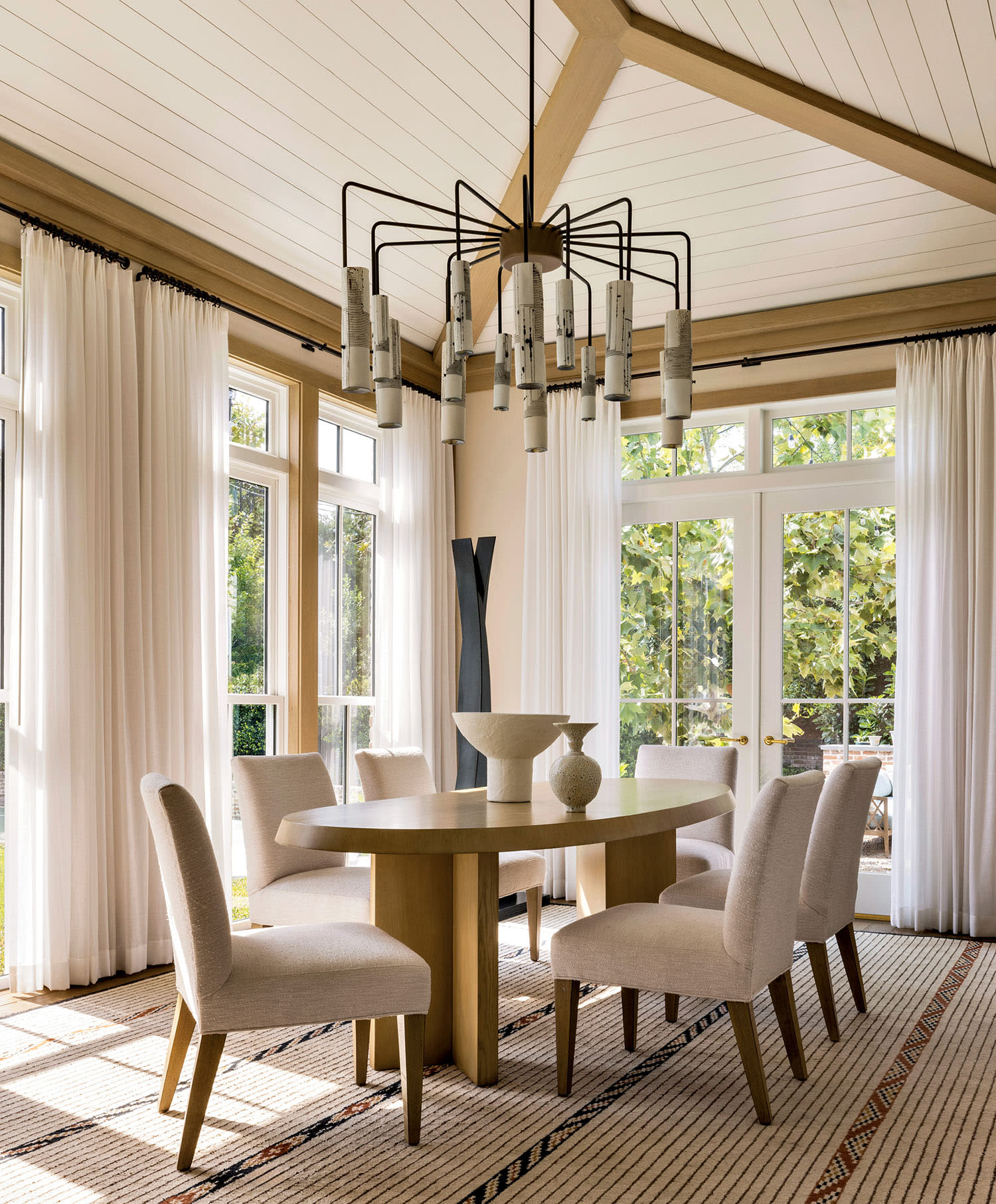 Elegant dining room with large windows, light wood ceiling beams, and a modern chandelier. Features a wooden table, six beige chairs, and sheer curtains.