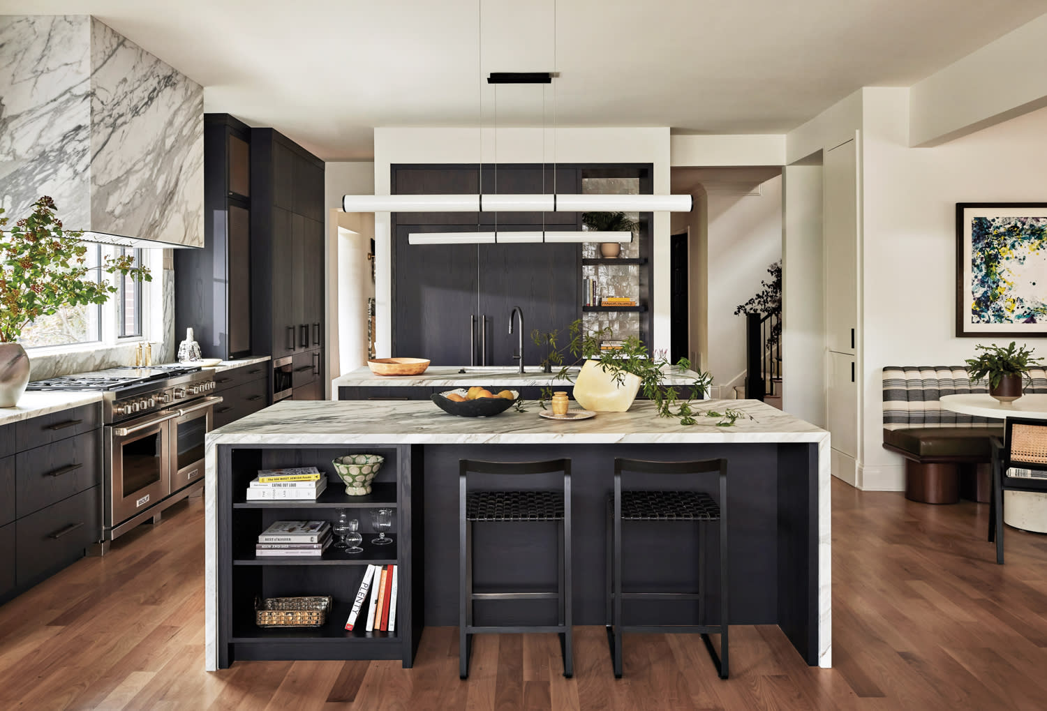 Modern kitchen with a marble island, black cabinetry, and wooden floors. Bar stools, potted plants, and abstract art add a stylish, inviting feel.