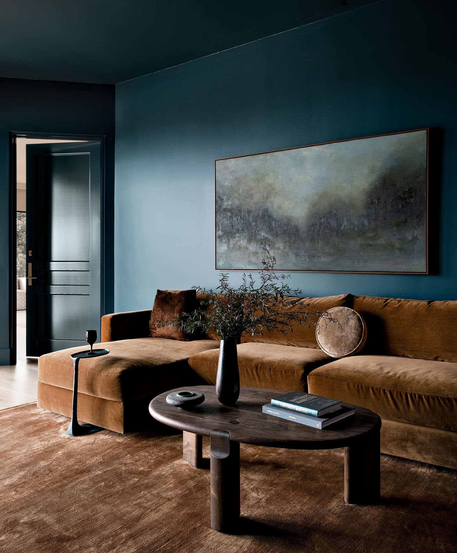 A brown couch sits against a blue wall with a landscape portrait hanging above and a nearby coffee table.