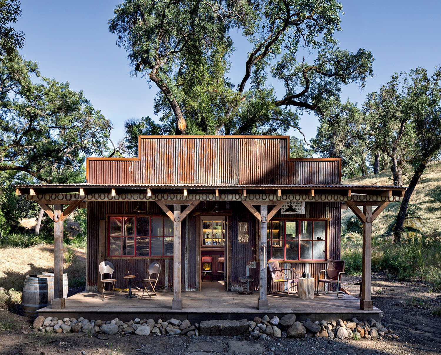 Rustic, weathered wooden cabin with a tin roof nestled among tall trees. Porch with chairs and a barrel, conveying a nostalgic, tranquil vibe.