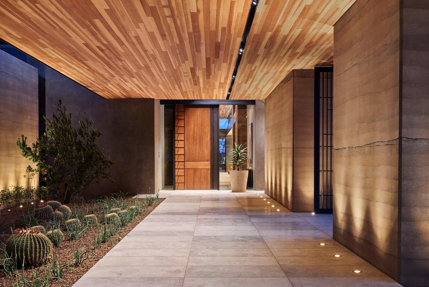 Modern entryway with warm wood ceiling, soft lighting, and stone walls. A path leads to a wooden door, bordered by desert plants and cacti, creating a serene atmosphere.