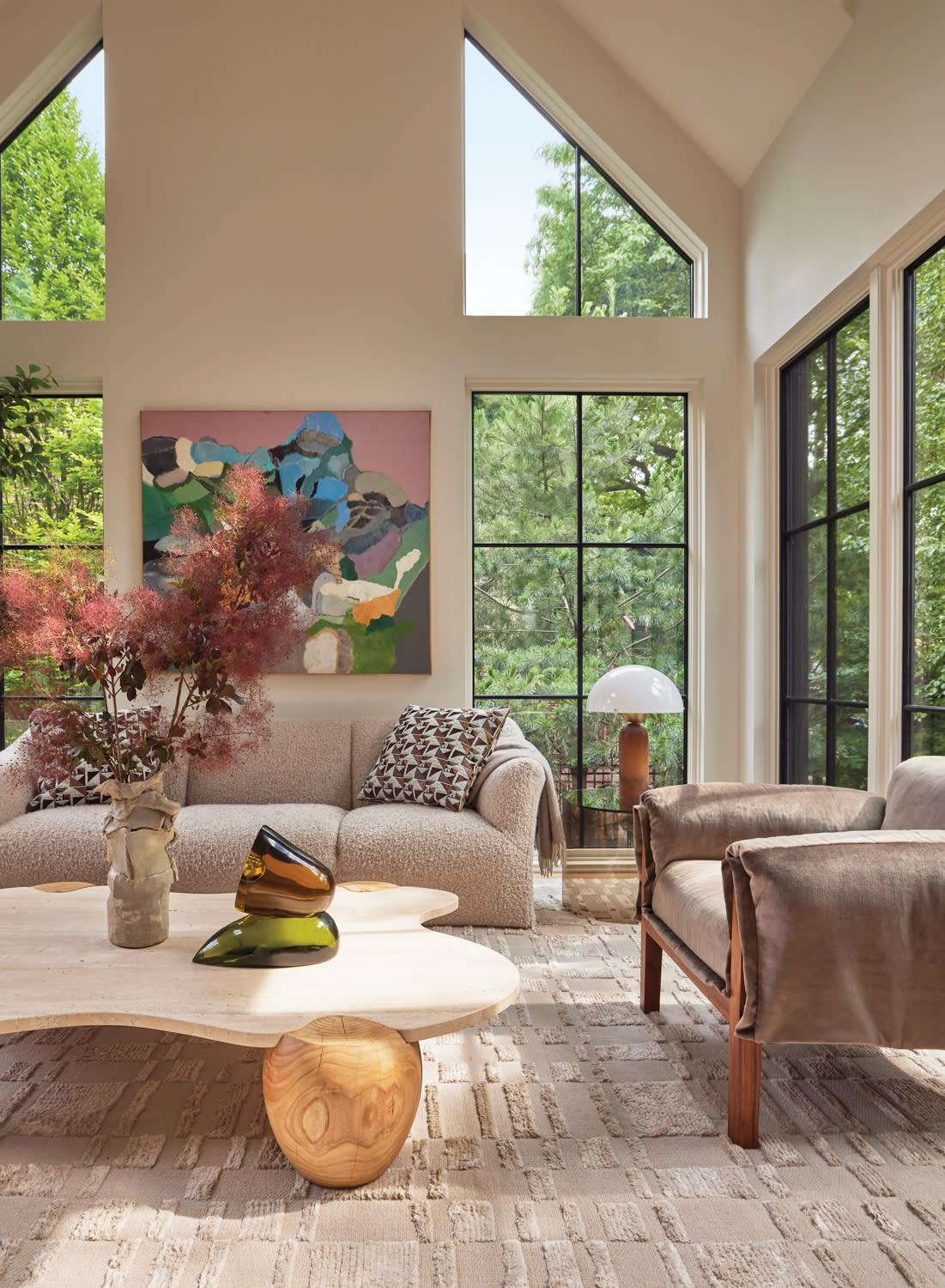 Bright modern living room with tall windows and natural light. Features beige sofa, abstract art, wood table, vase, and trees outside. Calm ambiance.