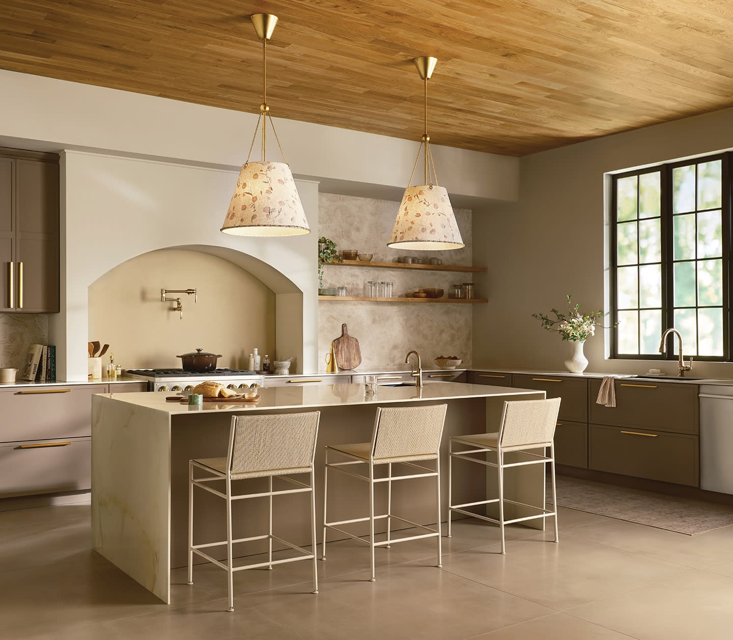 Warm, spacious kitchen with wooden ceiling, modern beige cabinetry, and marble island with four cushioned stools. Two ornate pendant lights hang above, creating a cozy ambiance. Natural light streams through a large window, enhancing the inviting atmosphere.