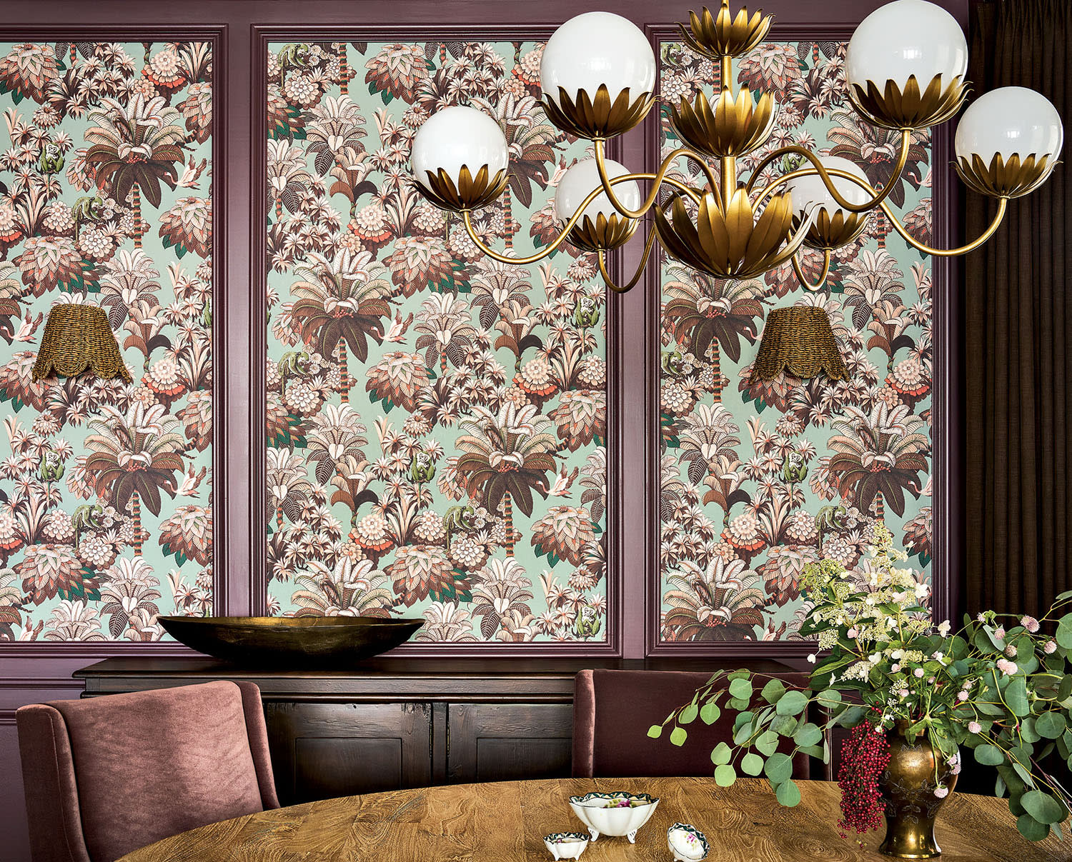 A wood dining table sits beneath an ornate chandelier in a room decorated with patterned wallpaper.