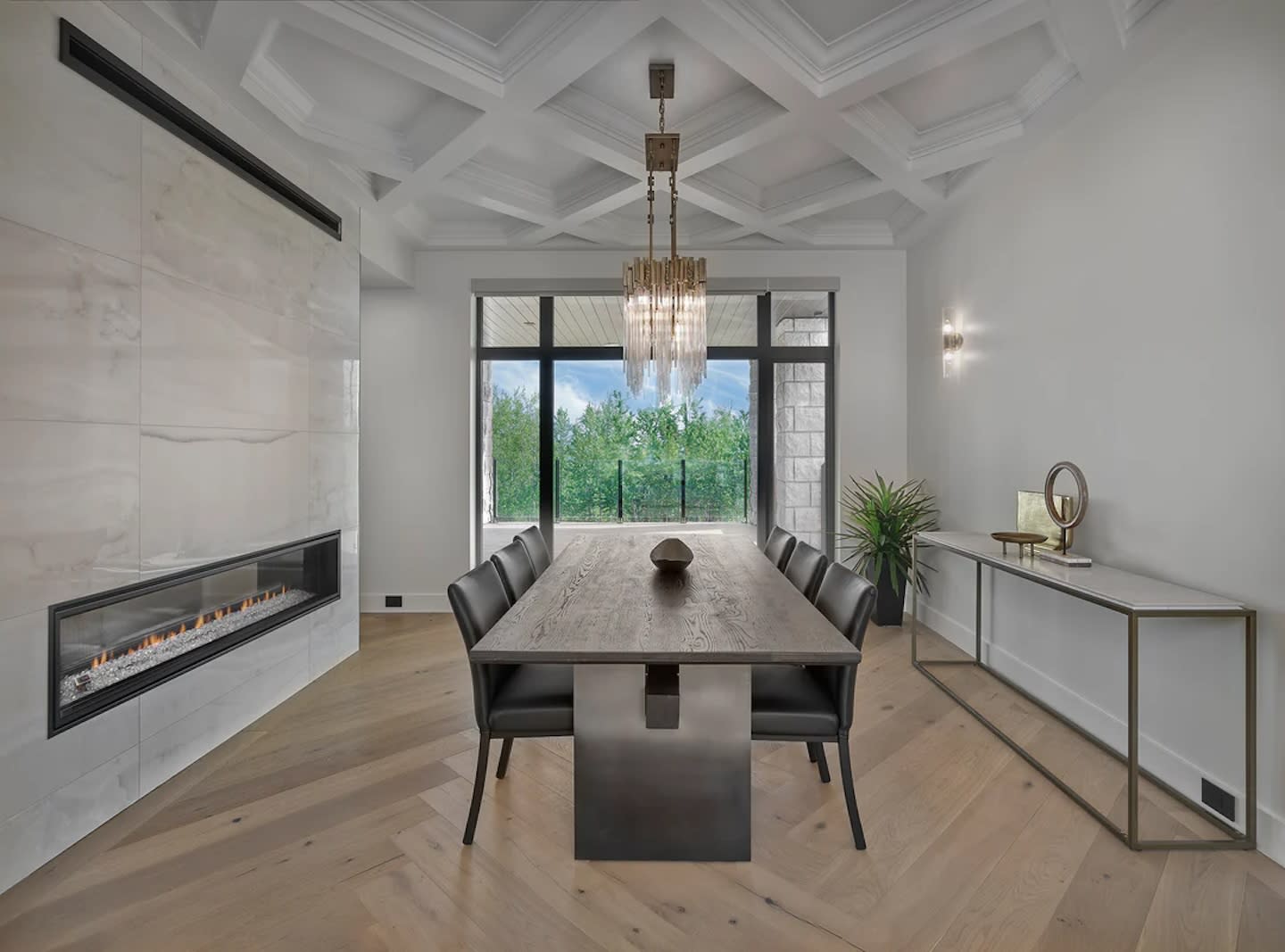 Modern dining room with a large wooden table, black chairs, and a chandelier. A large window shows a green outdoor view. Marble fireplace on the left.