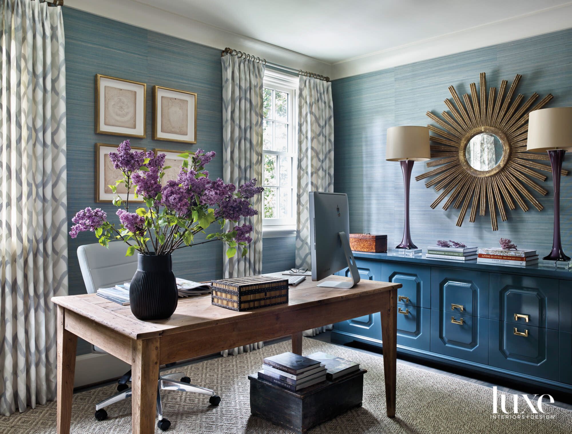 Home office with blue grasscloth wall covering, light-colored wood desk, lacquered blue cabinet, pair of table lamps and sunburst mirror