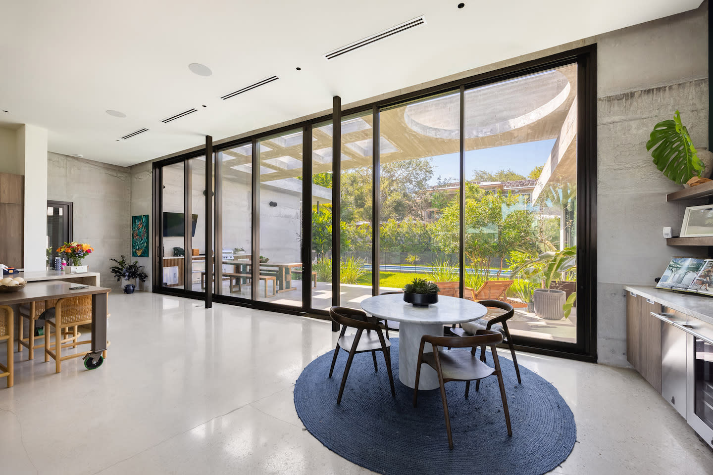 Sun-filled dining nook anchored by a round white-marble pedestal table and four curved wood chairs atop a navy braided rug on polished concrete floors. A sweeping wall of black-framed floor-to-ceiling glass sliders blurs the line between the minimalist interior’s raw concrete walls and the verdant patio garden outside.