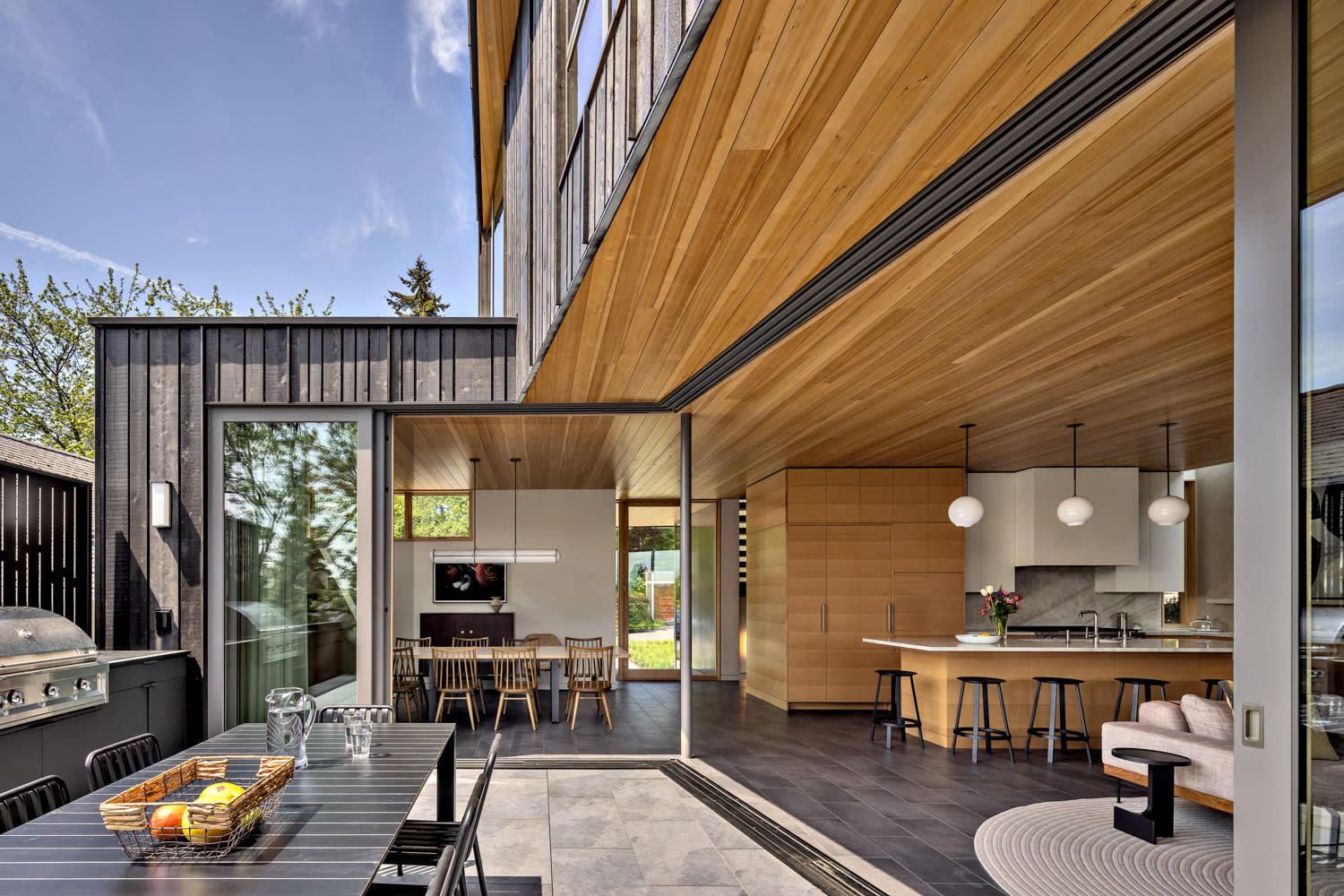 Modern open-concept patio and kitchen with wooden ceiling and black dining table creating a warm, inviting space.