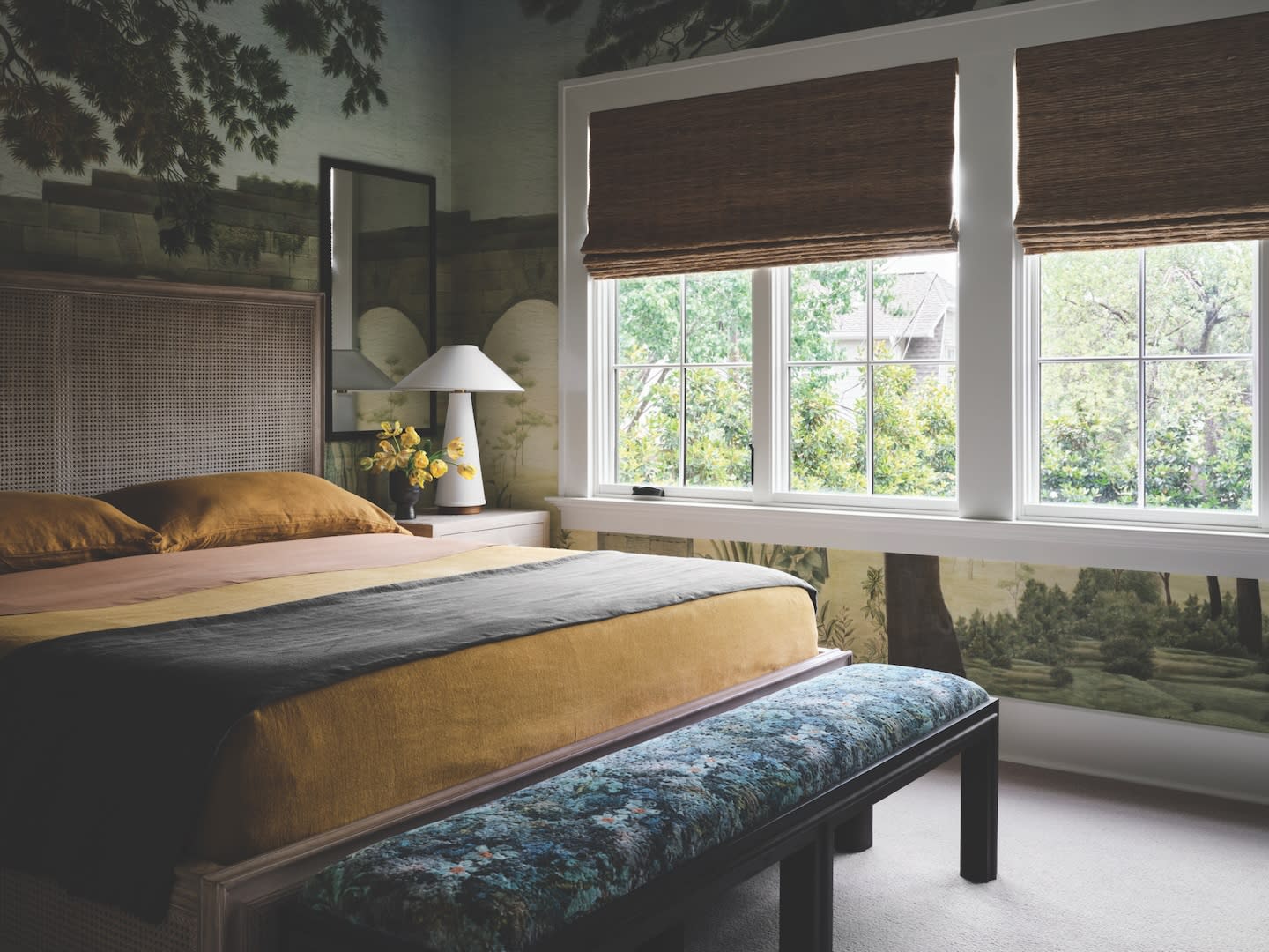 A cozy bedroom with nature-themed wallpaper and large windows with woven blinds. The bed has earthy-toned bedding and a floral bench at the foot.