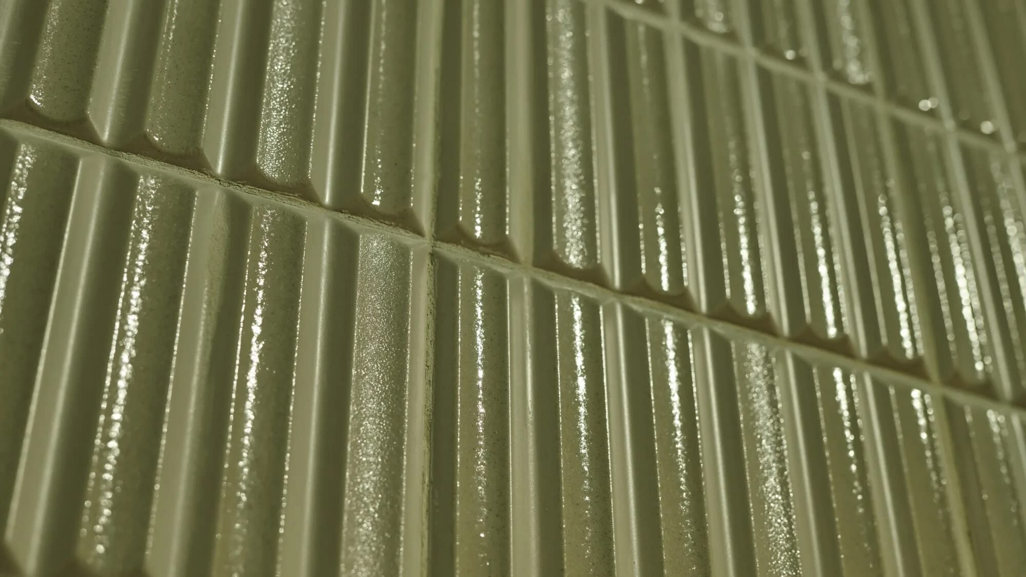 Close-up of a shiny, light green textured tile surface with parallel vertical ridges. The lighting highlights the glossy and reflective finish.