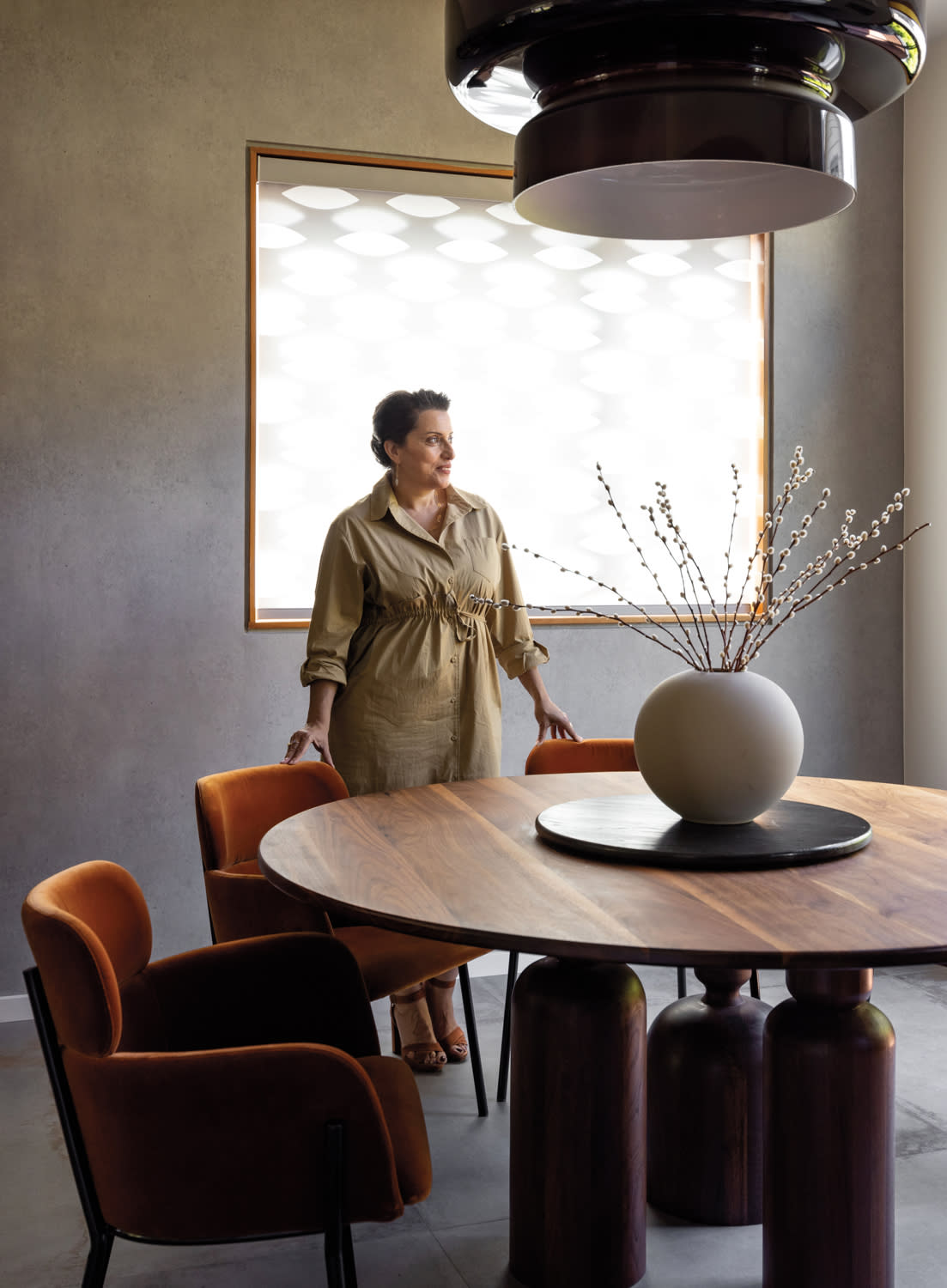 Designer Shoshi Cohen standing behind round wood table and rust-colored velvet-upholstered chairs.