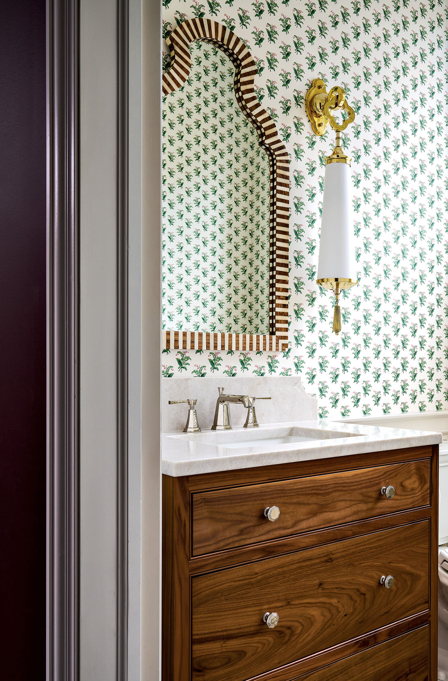 A bathroom decorated with floral wallpaper in white and green, with a sink, mirror and sconce.