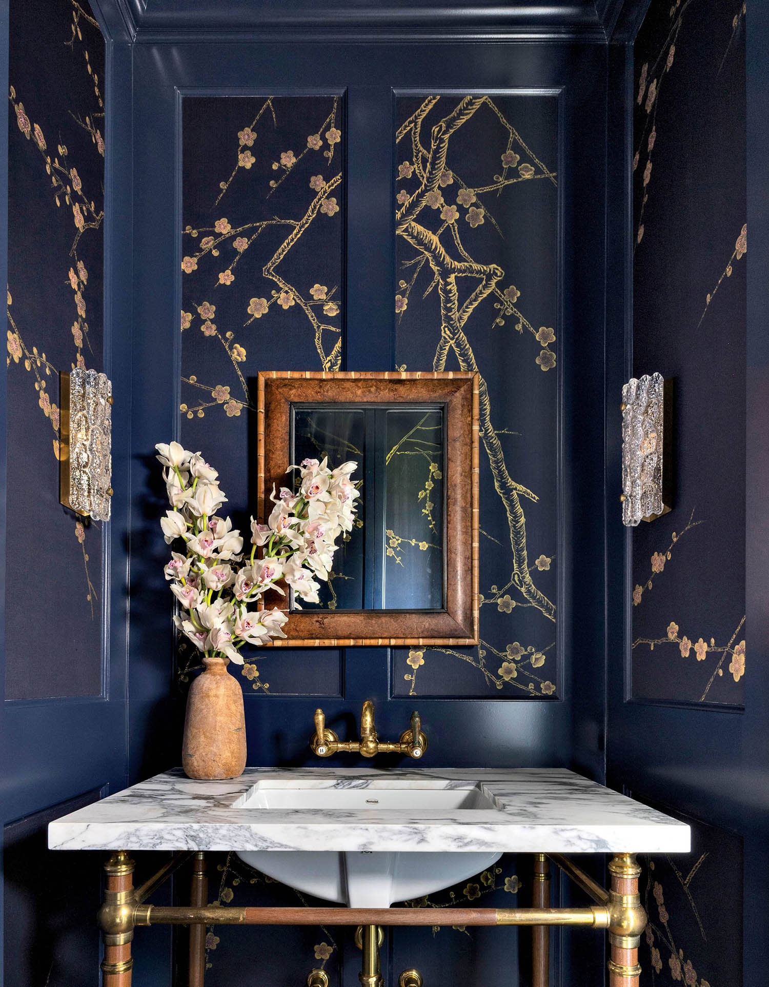 A bathroom with floral wall panelings and textured-glass sconces alongside a wood-framed mirror.