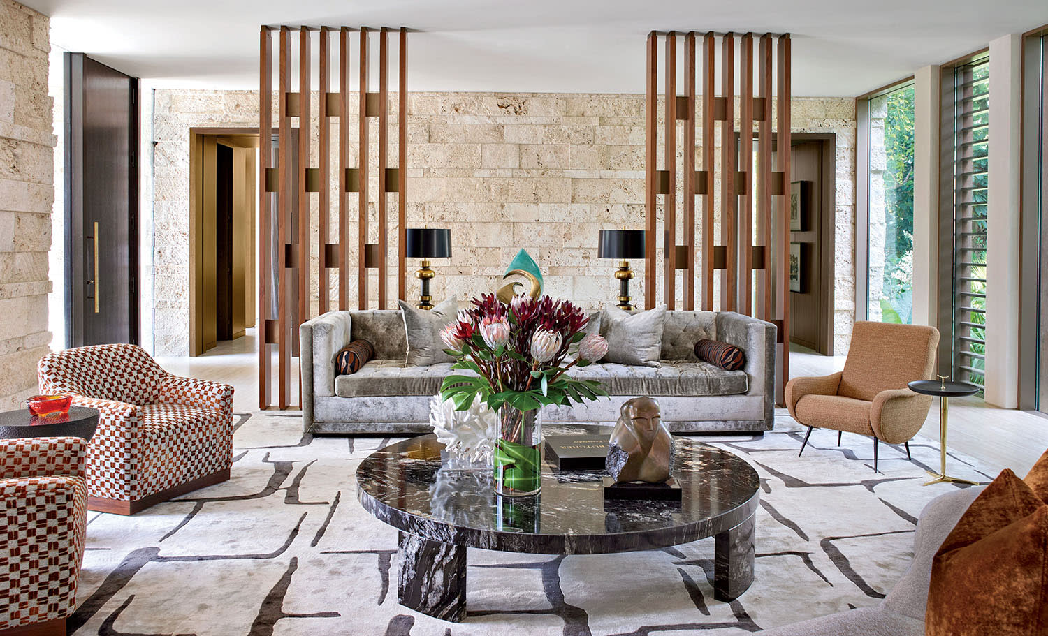 Modern living room with a marble coffee table, plush sofa, and patterned chairs. Natural stone walls are complemented by large windows and artistic decor.